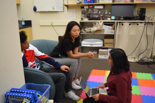 Three undergraduate members of the lab having a discussion. Two of them are seated on a couch, one of them is seated on a colorful play mat on the floor