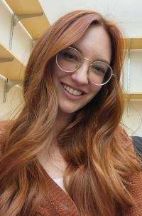 A selfie of Shannon. Shannon is wearing glasses with a gold frame and clear lenses, and shannon is wearing a redish orange v neck sweater