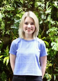 Annika is standing in front of a wall of leaves. Annika is wearing a blue and white check top with black pants.