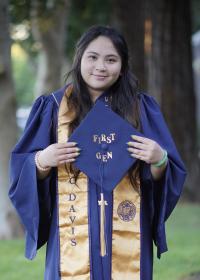 Van standing with her blue robe and gold graduation stole holding her graduation cap that has "first gen" written on it.