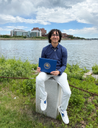 Smiling person seated by river holding blue diploma cover, campus across water