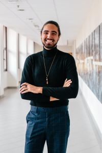 Erim standing in a hallway with his arms crossed. Erim is wearing a black turtleneck and blue slacks